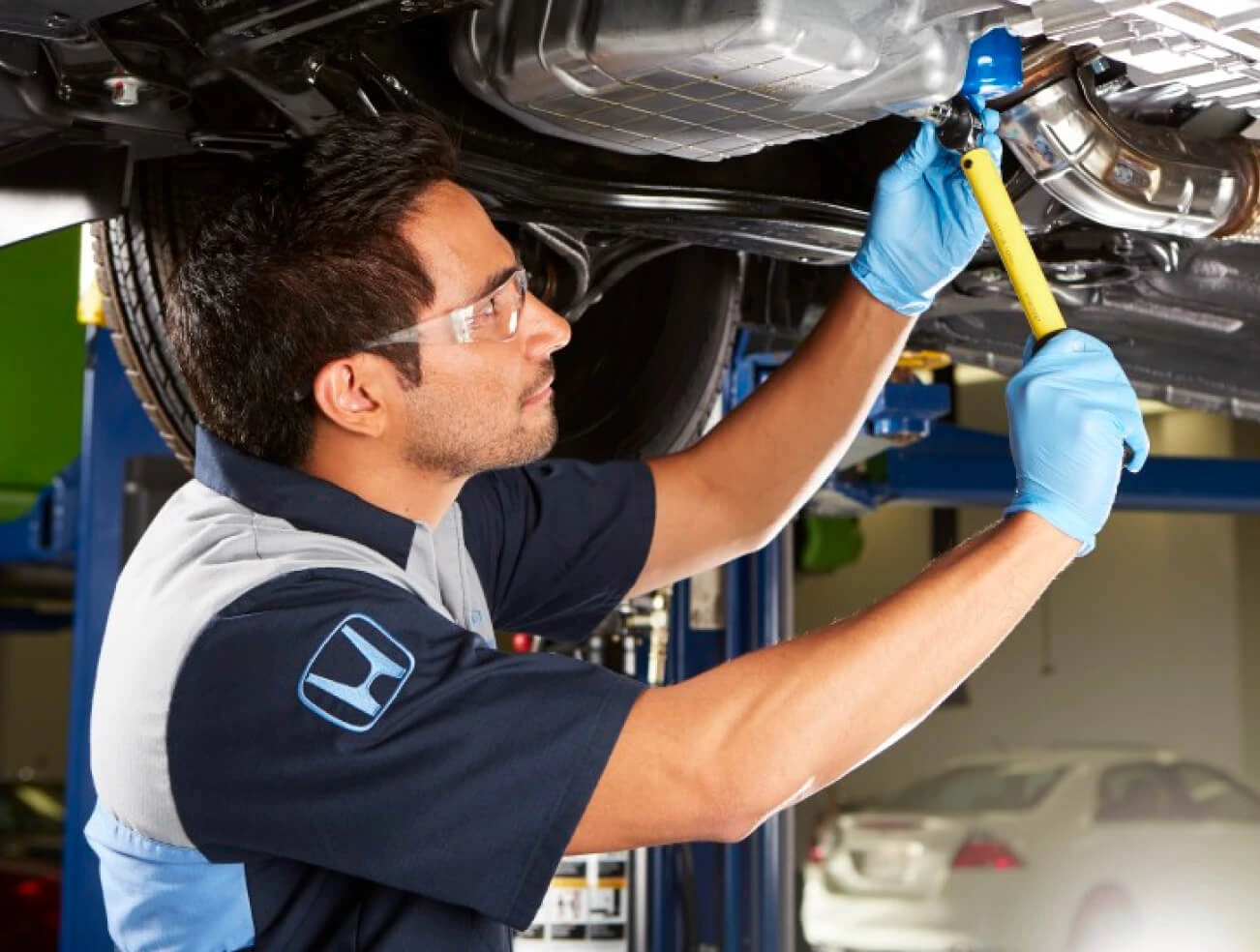 Honda technician performs underbody maintenance on a vehicle raised in a dealership service bay.