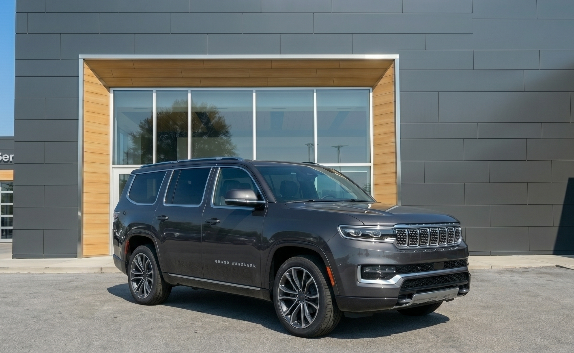 A dark gray Jeep Grand Wagoneer luxury full-size SUV parked on the lot in front of a modern dealership showroom facade with wood trim accents in Raleigh.