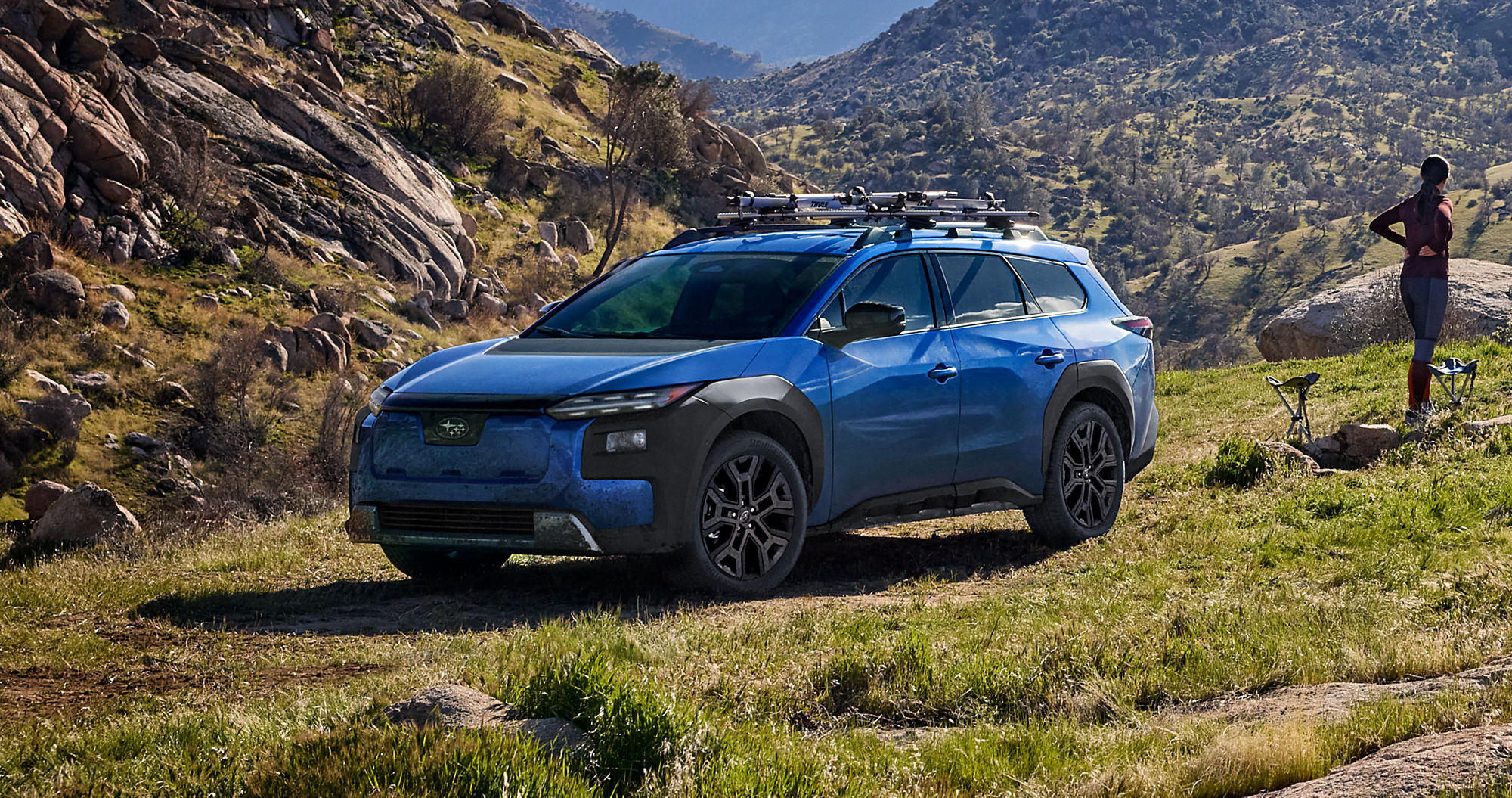 2027 Subaru Trailseeker electric SUV in blue parked on a grassy hillside with a roof rack and bikes