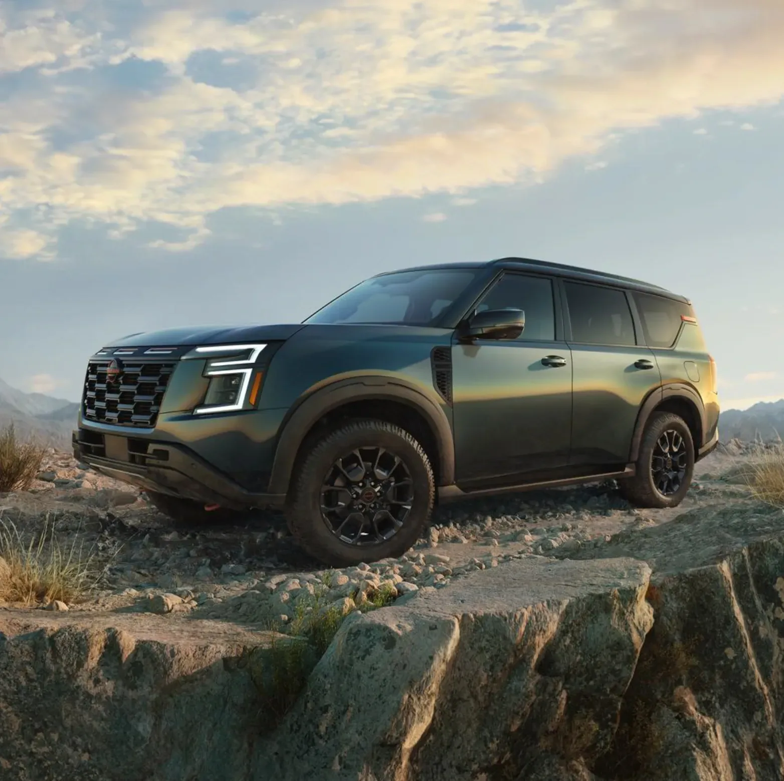 Dark green Nissan SUV with black wheels and a red tow hook parked on a rocky ridge under a cloudy sunset sky, mountains behind