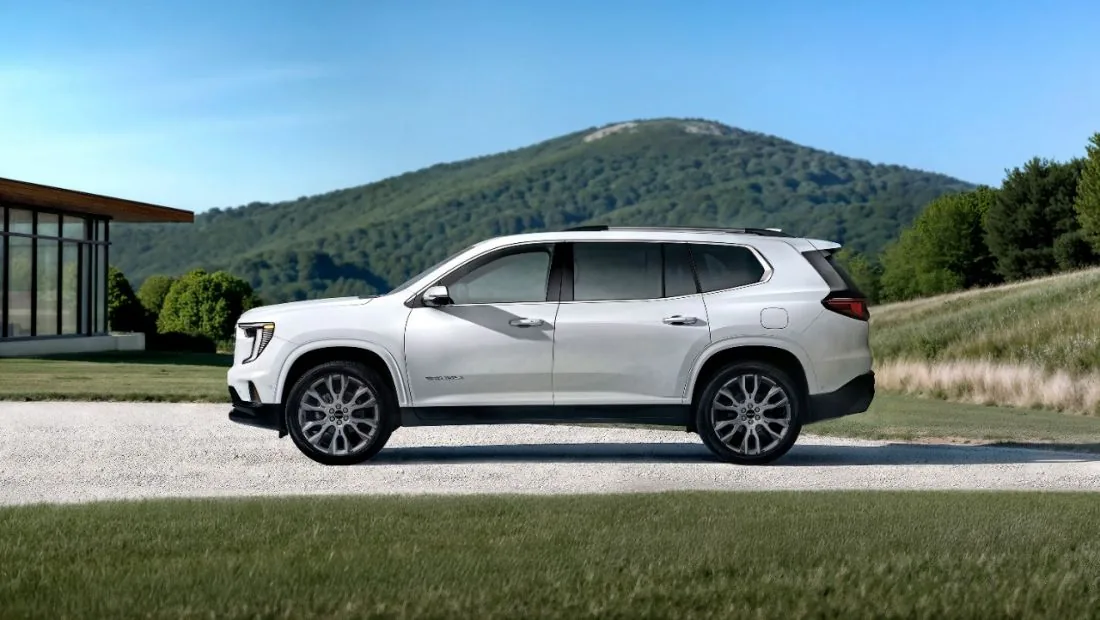 Side profile view of a white GMC SUV with a Denali badge, parked in front of a modern house with a mountain in the background