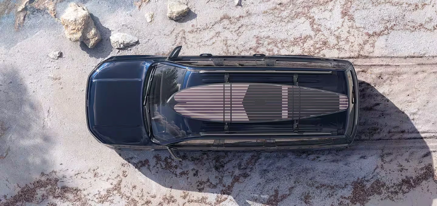 Overhead view of a dark blue 2027 Jeep Grand Wagoneer SUV with a striped surfboard strapped to its roof rails, parked on pale sandy ground with scattered rocks and a long shadow