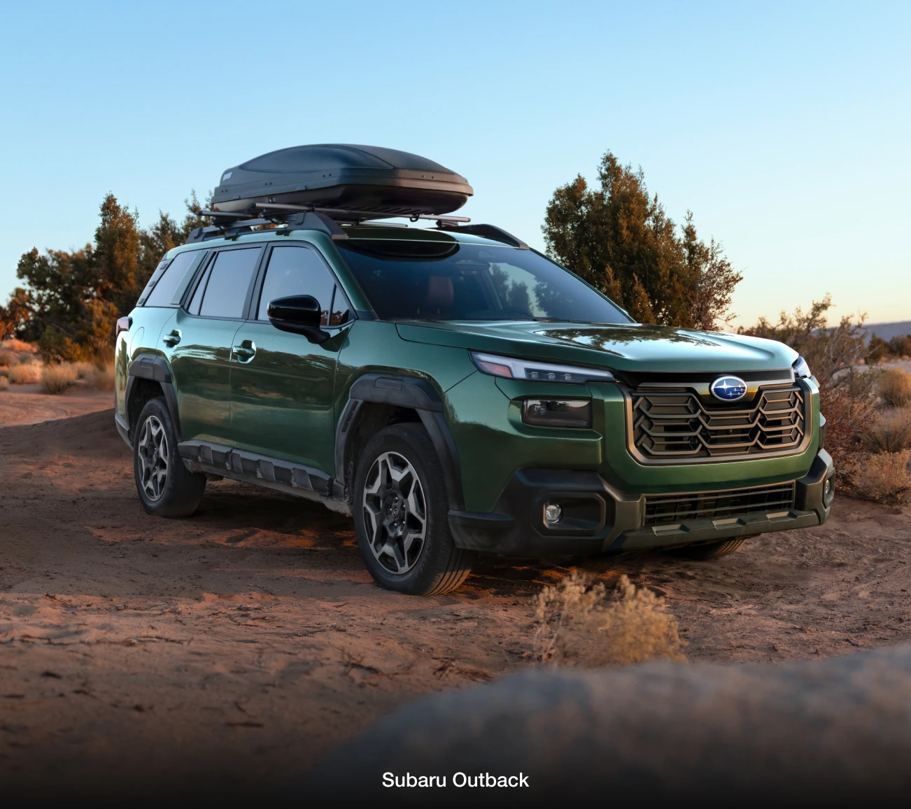 A green Subaru Outback with a roof cargo box parked on a sandy desert trail.