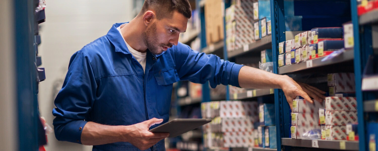 A parts department employee checks boxed automotive parts on a shelf using a tablet at a dealership.