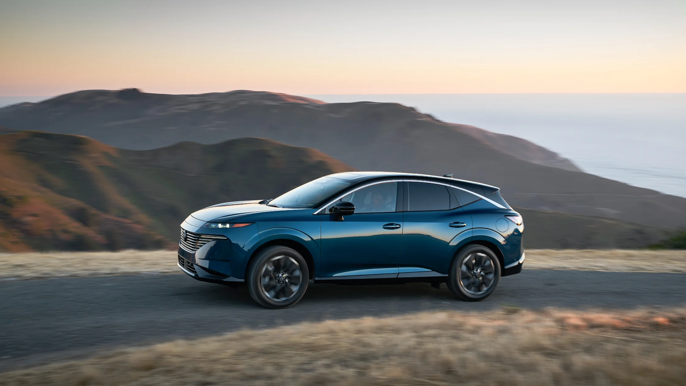2026 Nissan Murano® blue SUV side profile driving along coastal hills at sunset with ocean view