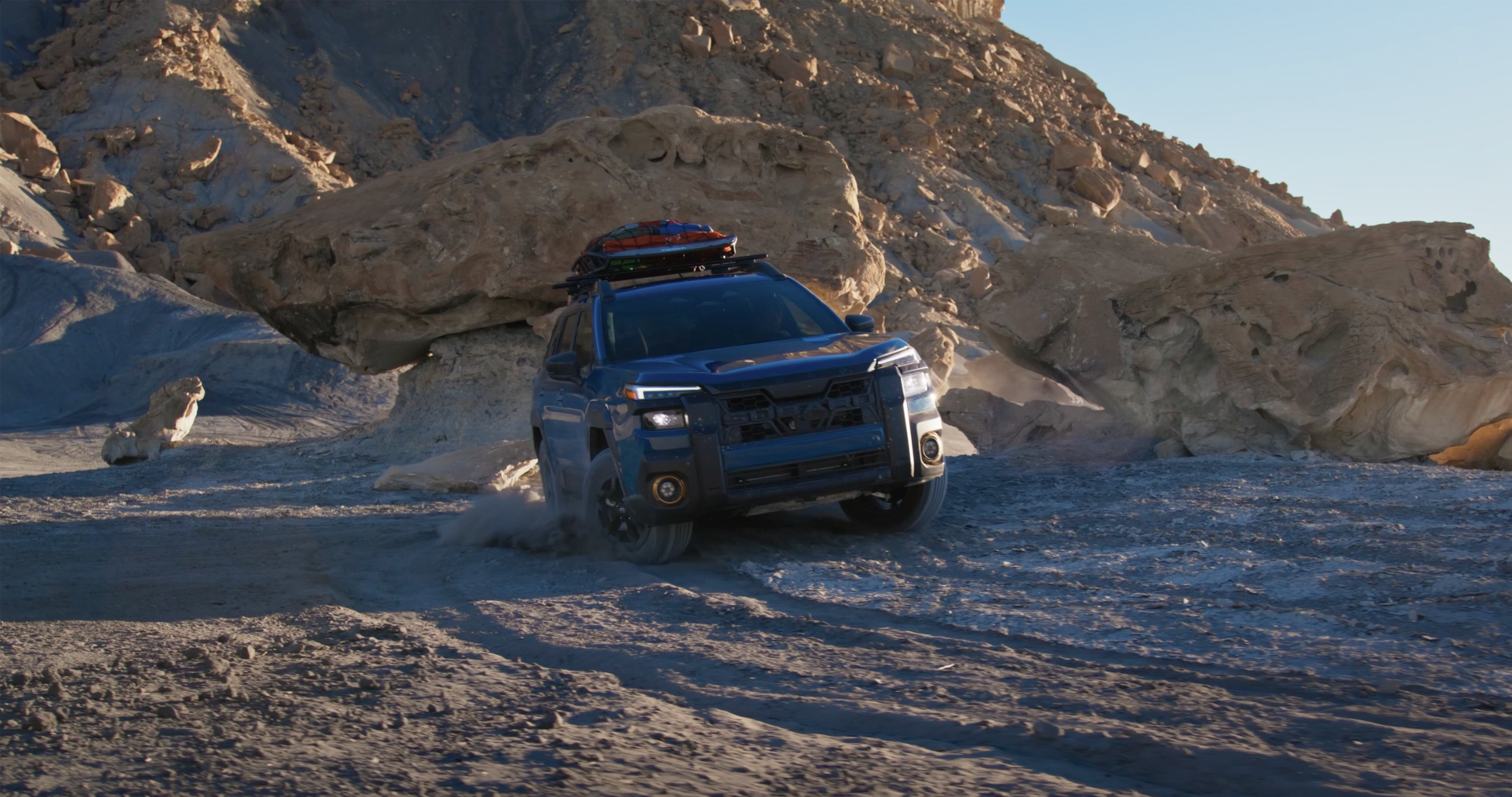 A geyser blue 2026 Subaru Outback Wilderness AWD SUV driving through various features of a desert biome..png / A 2026 Subaru Outback in emerald green and a 2026 Subaru Outback Wilderness in geyser blue parked next to each other on a mountain overlooking a desert. 