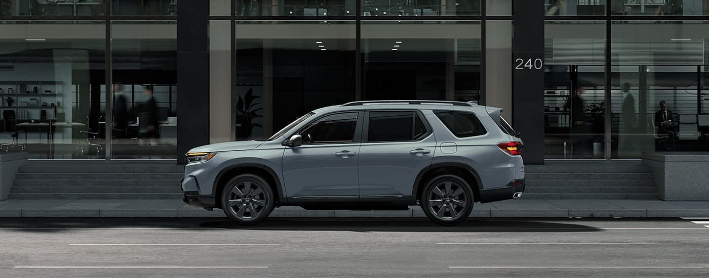 A grey 2023 Honda Pilot is shown from the side driving on a city street in front of a building with large windows.