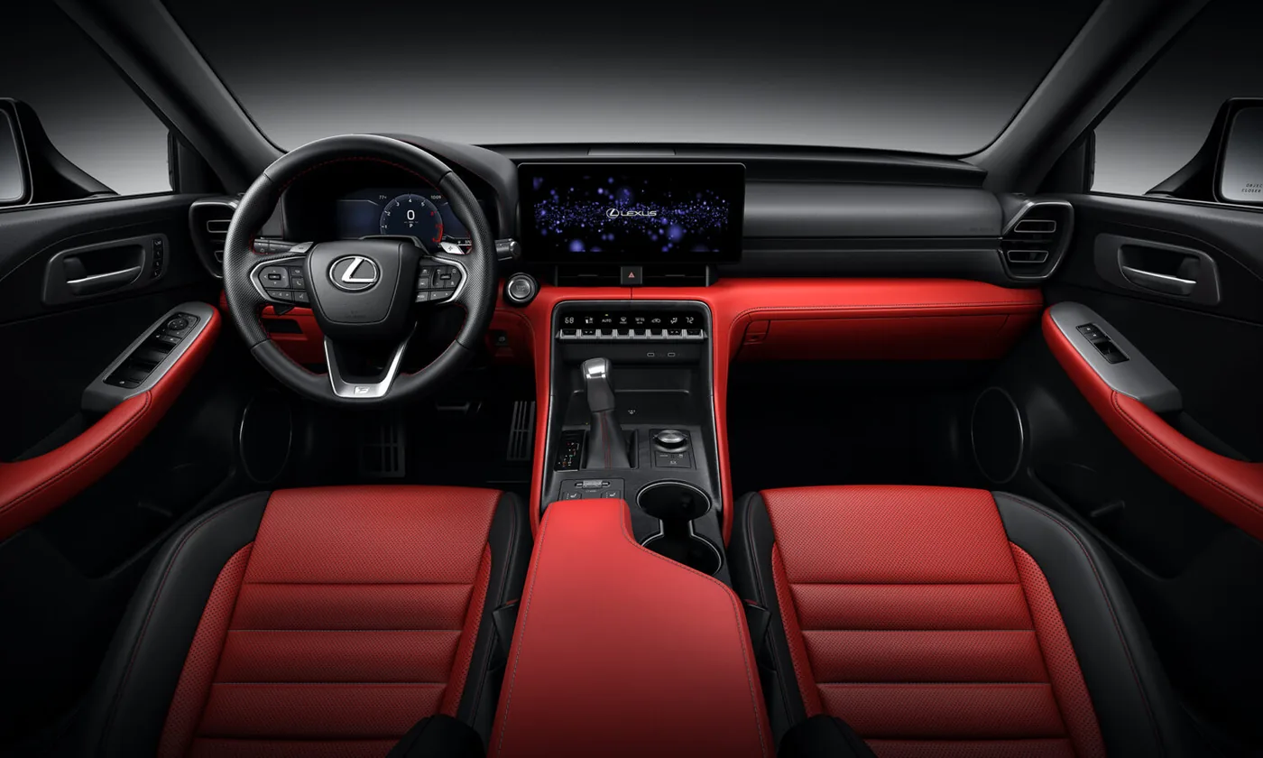Overhead studio view of a Lexus front cabin with red perforated leather seats and center armrest, red dashboard and door panels, a black steering wheel with the Lexus emblem, a wide touchscreen showing the Lexus logo, and a floor-mounted gear shifter.