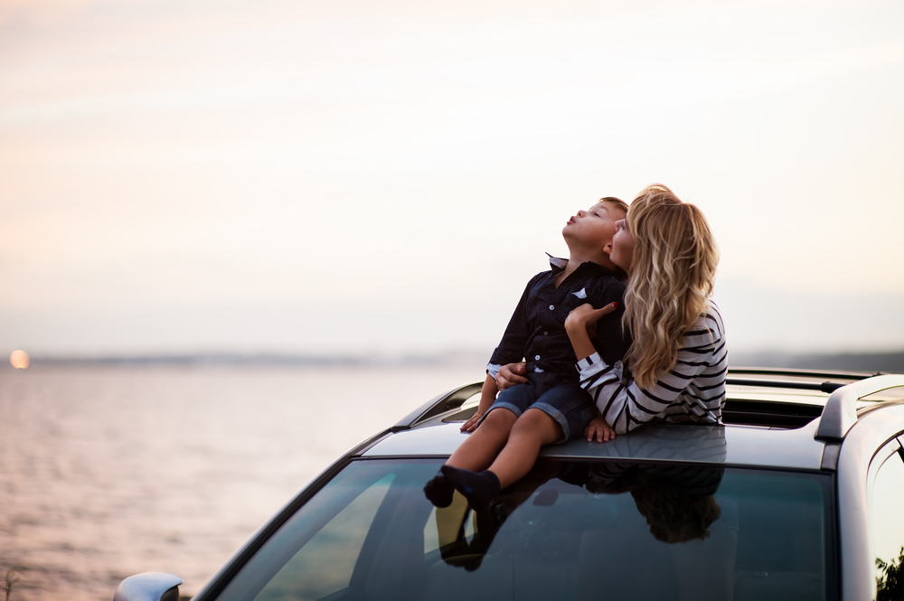 Mother standing through the moonroof of her car, while holding her son on the roof of car, as they look to the sky