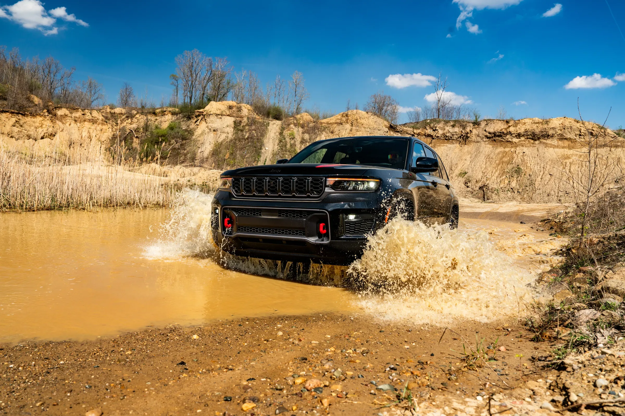 Dark gray 2027 Jeep Grand Cherokee L splashing through a muddy water crossing on an off-road trail, with red tow hooks and sandy cliffs in the background