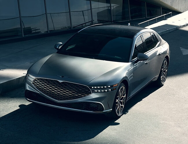 A silver 2025 Genesis G90 sedan with a diamond-pattern crest grille parked beside a modern glass building.