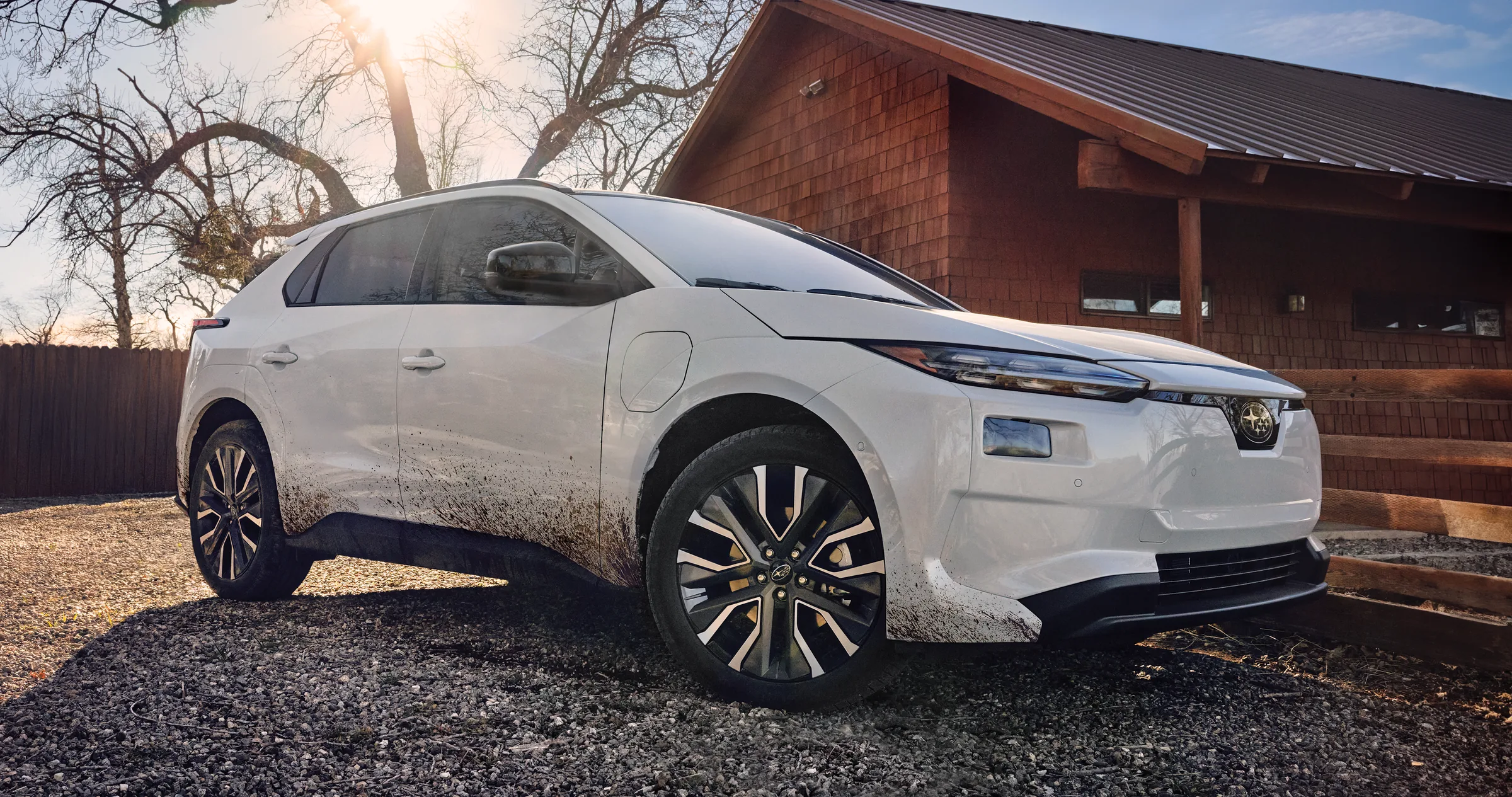 A static image of a white 2026 Subaru Solterra parked on a gravel surface beside a wooden building.