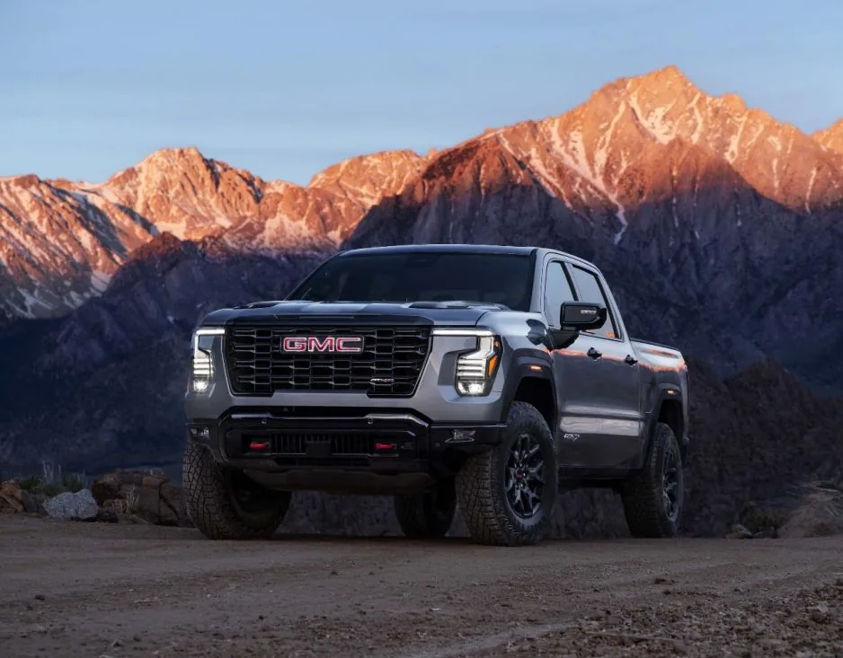 Front three-quarter view of a silver GMC pickup truck with off-road styling — round LED fog lights, black fender flares, and tow hooks — parked at a scenic overlook as mountains glow at dusk