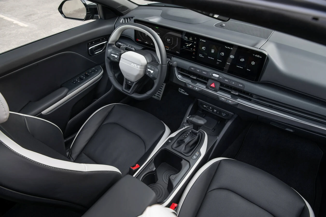 Interior of a 2025 Kia K4 showing the steering wheel with Kia badge, digital instrument cluster, widescreen infotainment display with home screen widgets, and two-tone black-and-white leather seats.