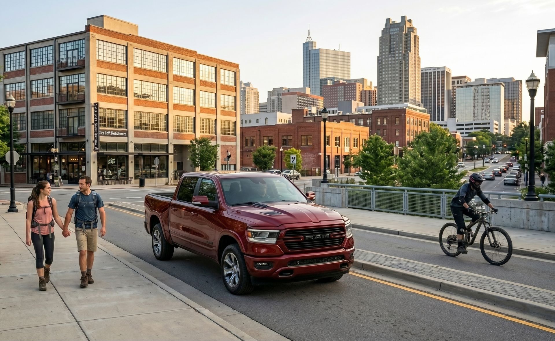 A dark red Ram 1500 crew cab pickup truck driving through a downtown city street with pedestrian hikers walking on the sidewalk and a cyclist nearby, featuring the illuminated downtown Raleigh skyline in the background.