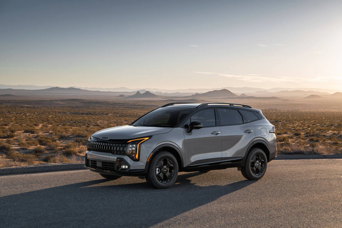Light gray 2027 Kia Sportage with black off-road-style wheels and roof rails, parked on a desert road at sunset with scrubland and distant hills.