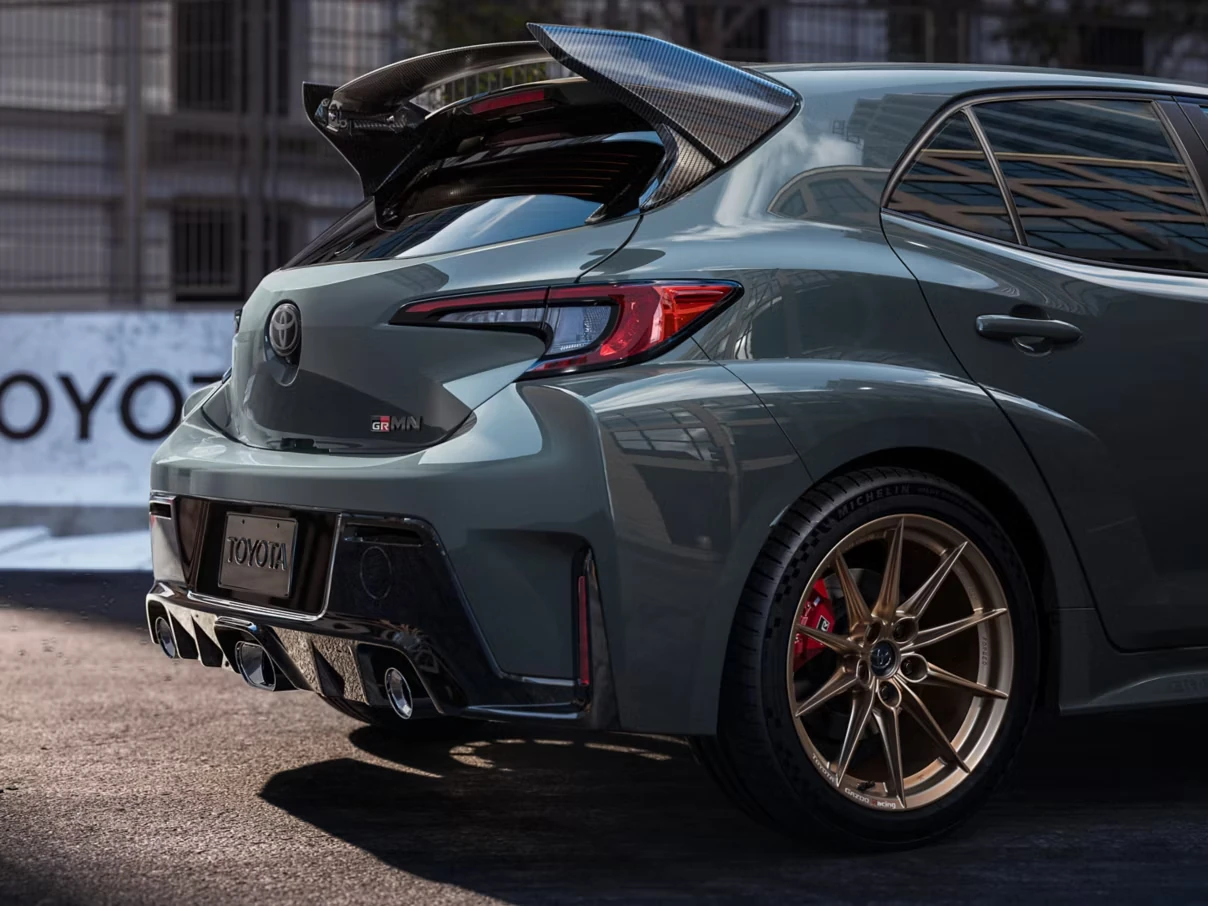 Rear three-quarter detail of a gray-green 2026 Toyota GR Corolla with a large carbon-fiber rear wing, gold multi-spoke wheels, red brake calipers and chrome exhaust tips, parked on a city street