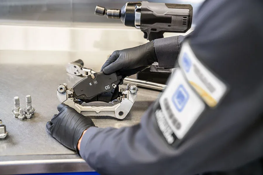 Female technician wearing safety glasses holds an ACDelco AGM battery near an open hood, preparing to install it, with equipment panels visible in the background