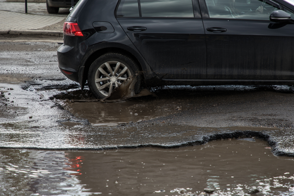 A black car is driving through a battered street with a large pothole, the cars back left tire is going through the pothole and it is raining and the streets look very wet