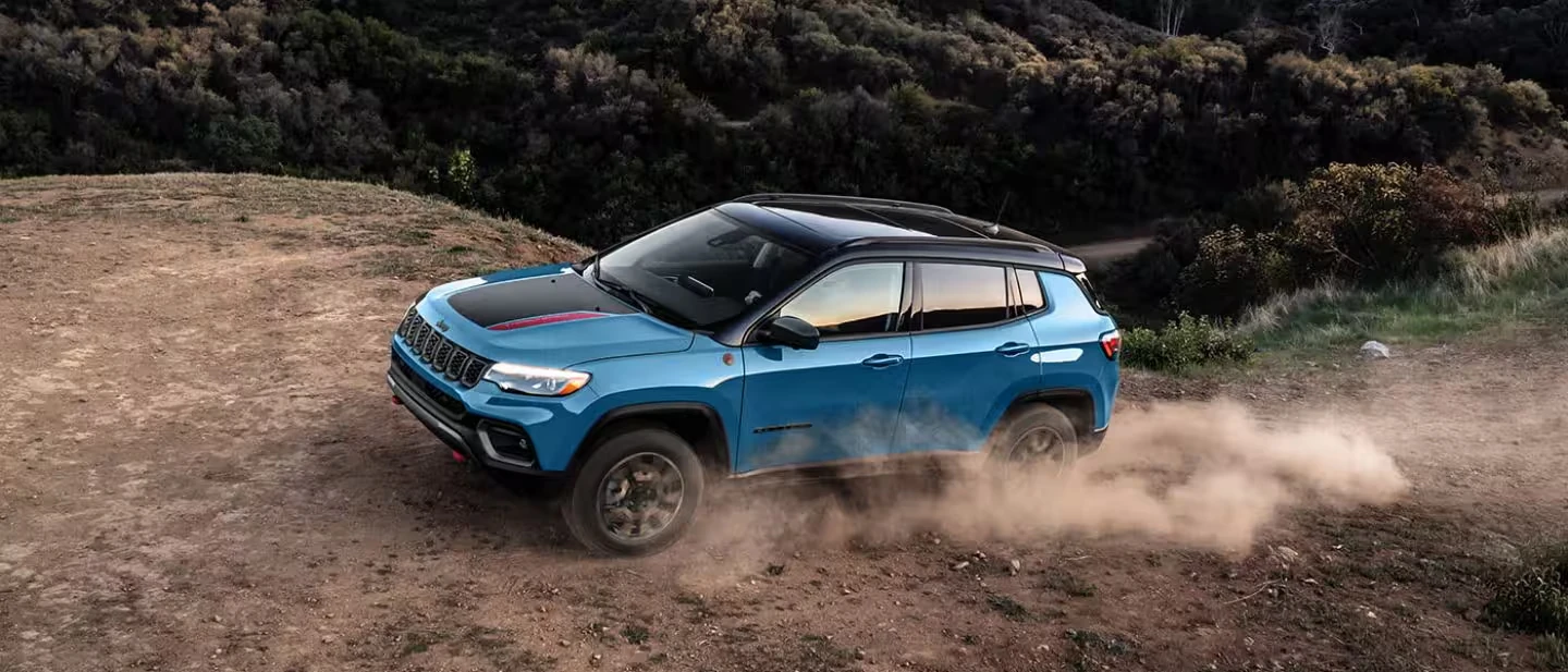 Blue 2026 Jeep Compass SUV with a black roof and a black hood decal with a red stripe, driving up a dirt hillside and kicking up a cloud of dust, with shrubs on the slope behind it