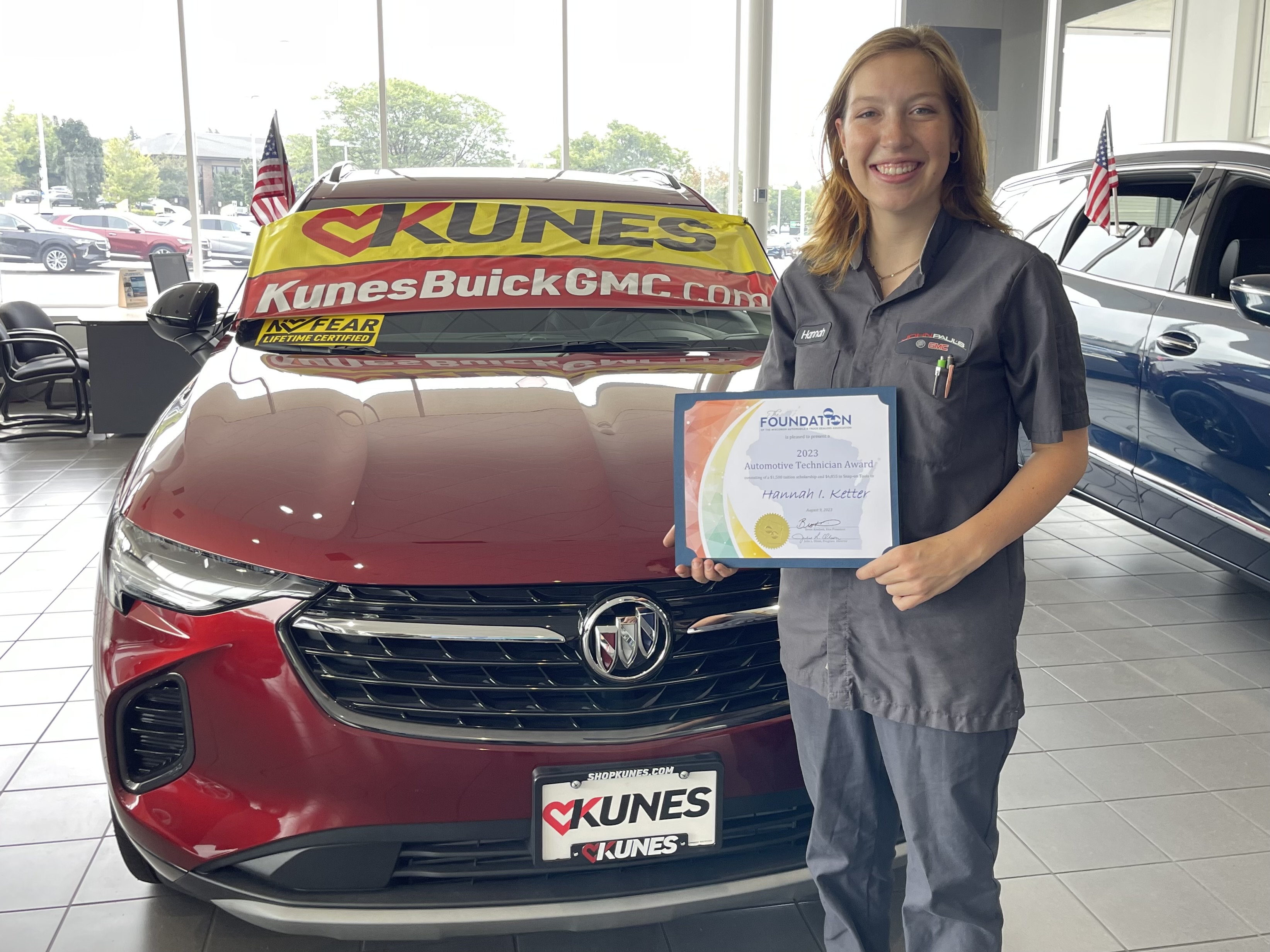 Hannah Ketter holding her scholarship in front of a new red Buick on the showroom floor at Kunes Buick GMC of Greenfield