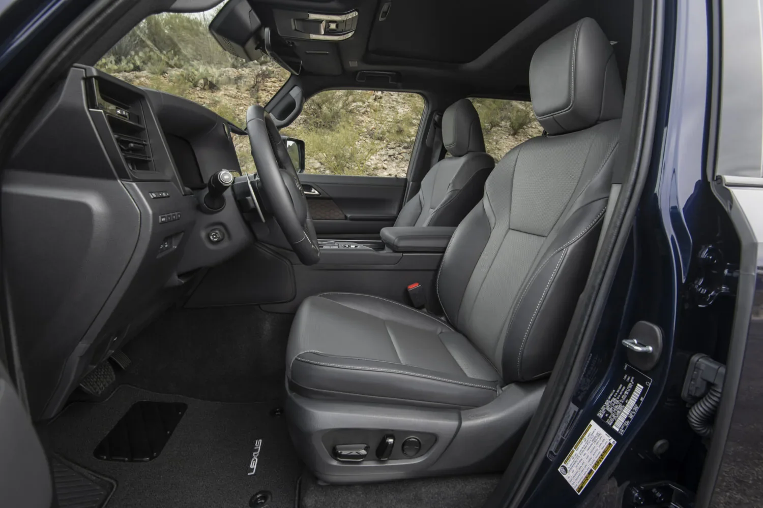 Driver-side interior of a Lexus SUV with the dark blue door open, showing a black leather front seat with gray stitching, power seat controls, a leather steering wheel, a black floor mat embroidered "LEXUS," and a rocky desert hillside outside the windows.
