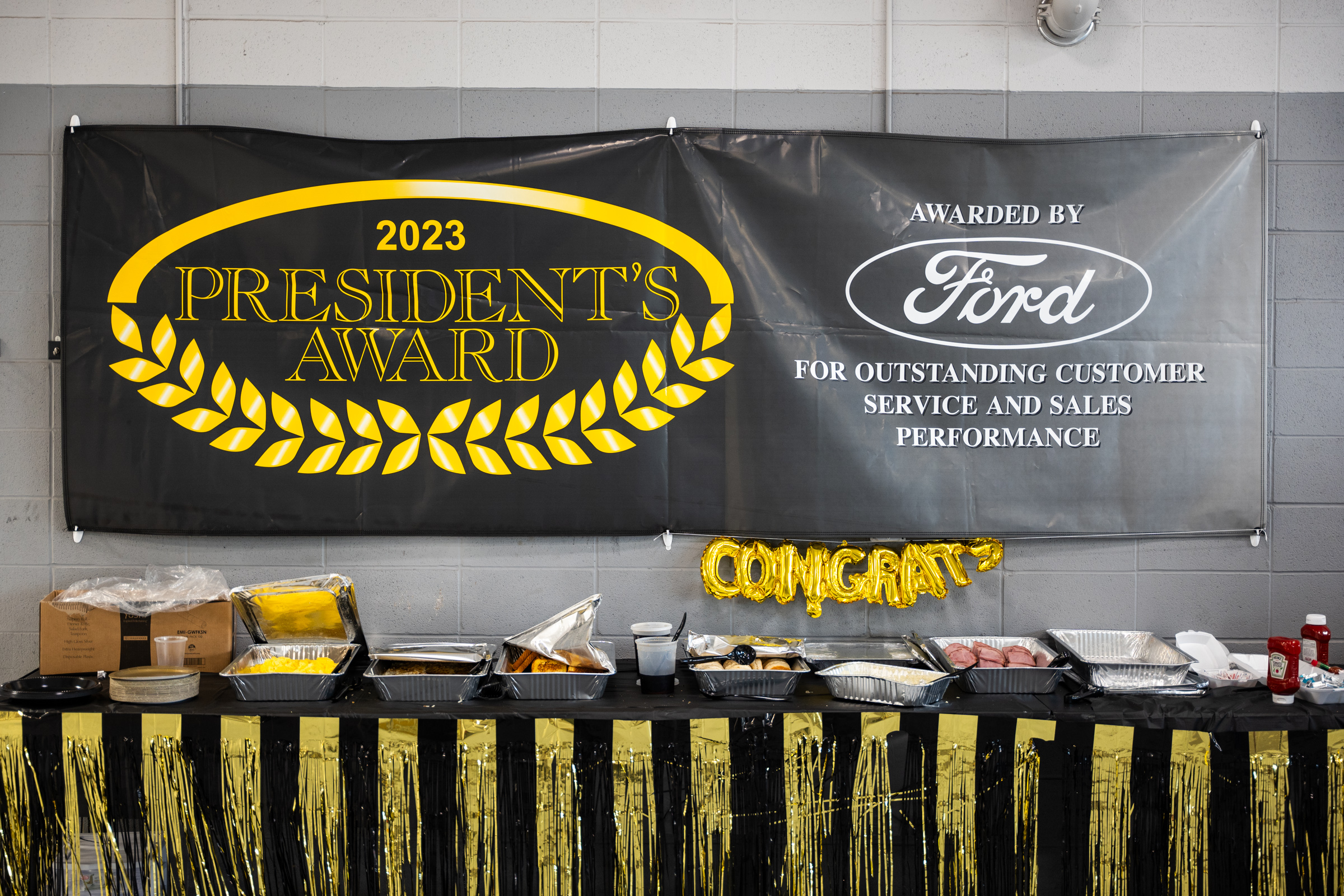 Banner hangs above a table of assorted foods. The banner reads: 2023 President's Award; Awarded by Ford for outstanding customer service and sales performance