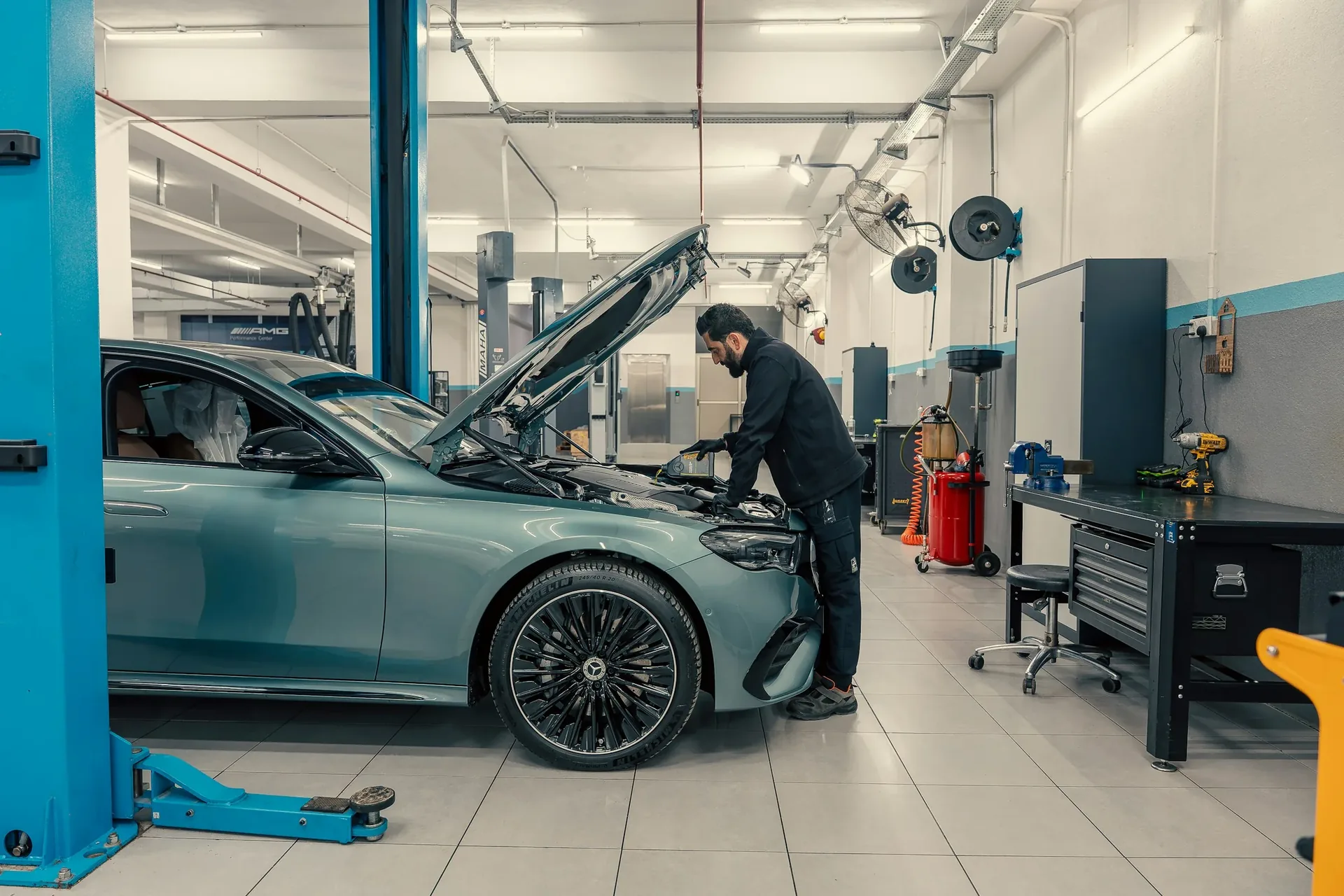 Mechanic wearing a Mercedes-Benz tri-star jacket working beneath a teal sedan raised on a service lift, with AMG Performance Center signage visible in the background