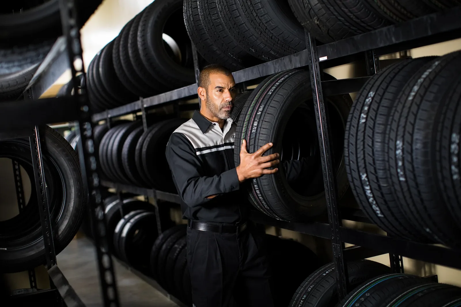 A technician in a gray and black service uniform holds a tire on a tall black storage rack, with rows of stacked tires on the shelves around him.