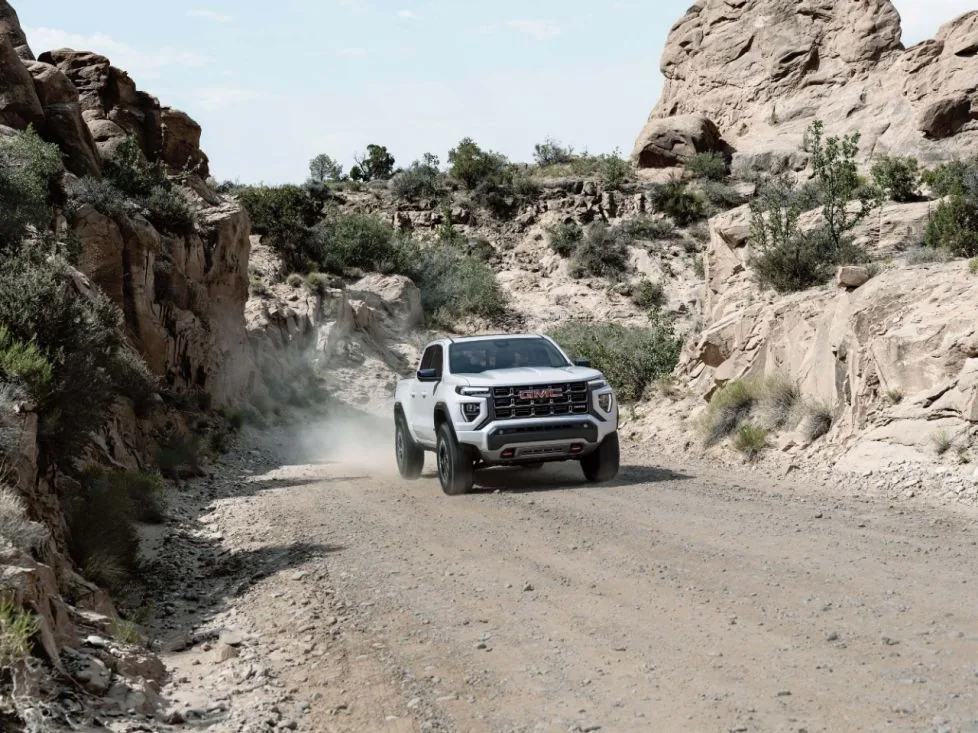 A white GMC pickup truck drives along a dirt road through a narrow desert canyon, with rock walls and sparse vegetation on either side