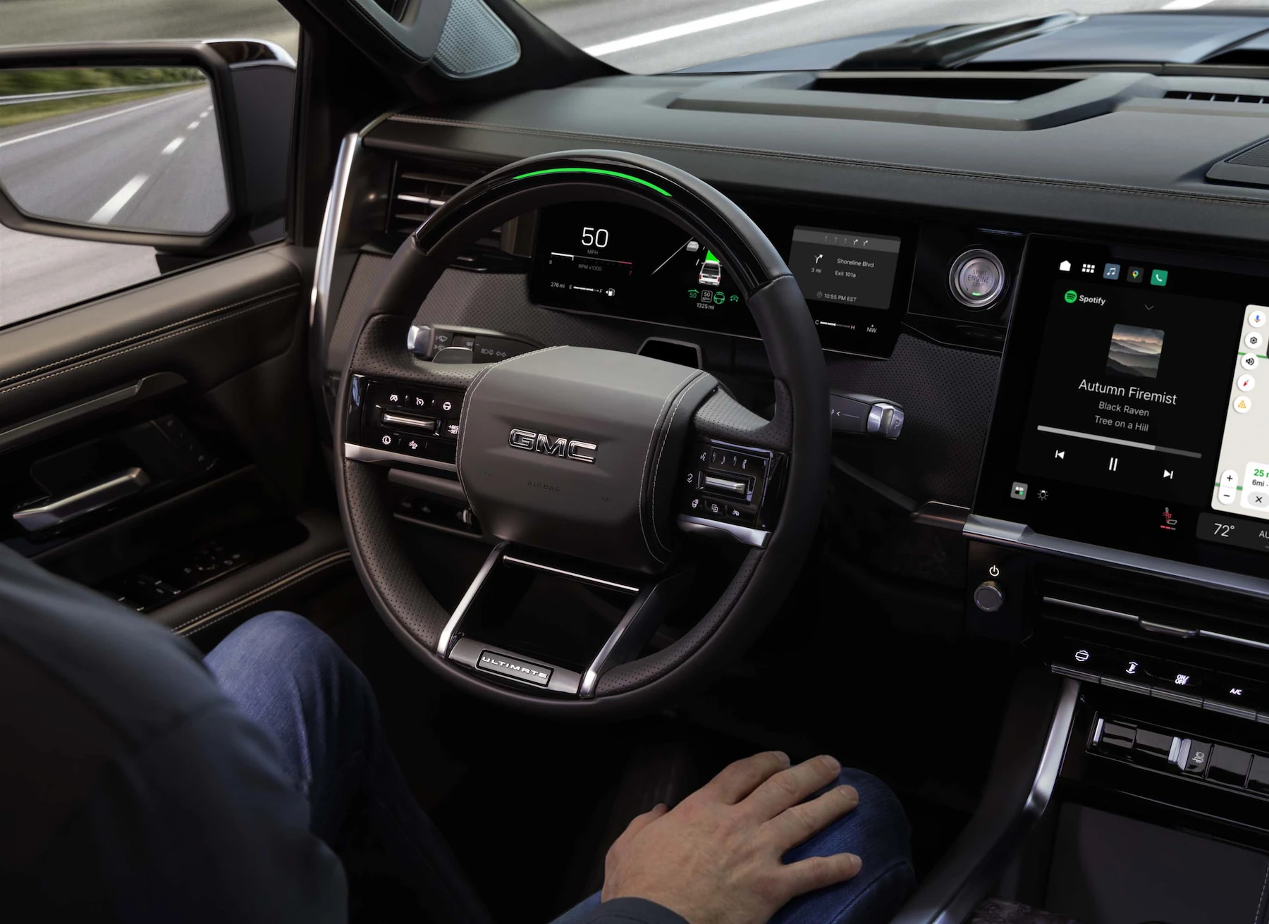 Over-the-shoulder interior view of a 2027 GMC Sierra 1500 Denali Ultimate in Stealth Black, showing Super Cruise hands-free driving engaged (green steering wheel light), 50 mph on the display, turn-by-turn navigation, and Spotify playing on the center touchscreen