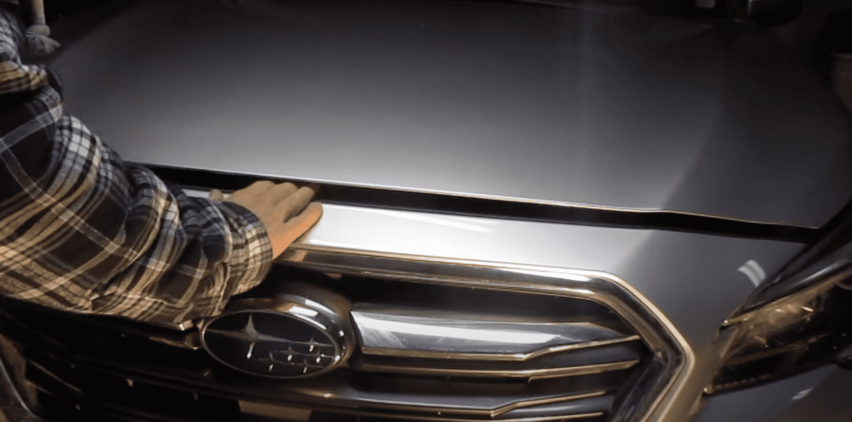 Hand reaching for the hood latch above a Subaru grille