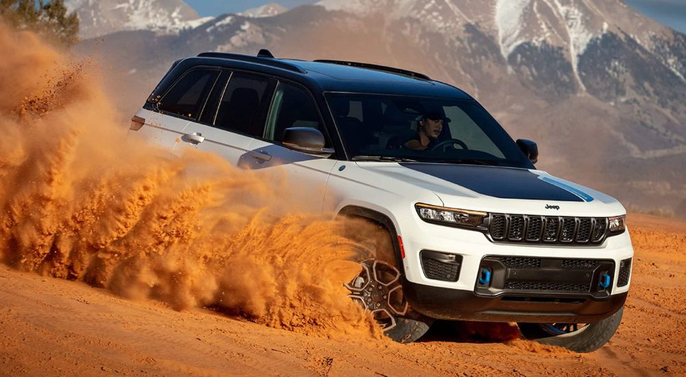 A white 2024 Jeep Grand Cherokee 4xe is shown from the front at an angle while driving through sand.