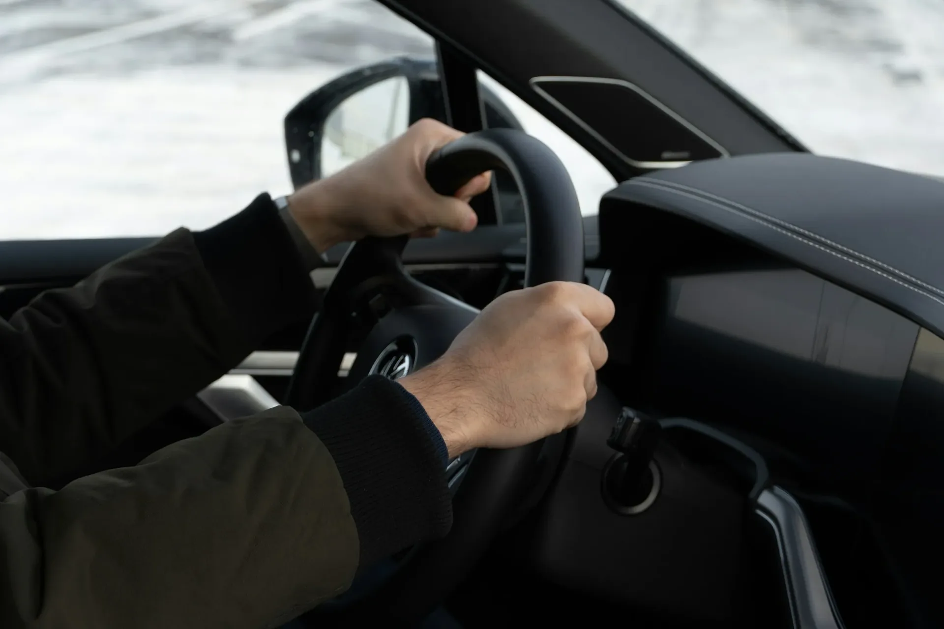 Driver's hands gripping a Volkswagen steering wheel with the VW logo on the hub, snow-covered surroundings visible through the windshield and a touchscreen display on the dash