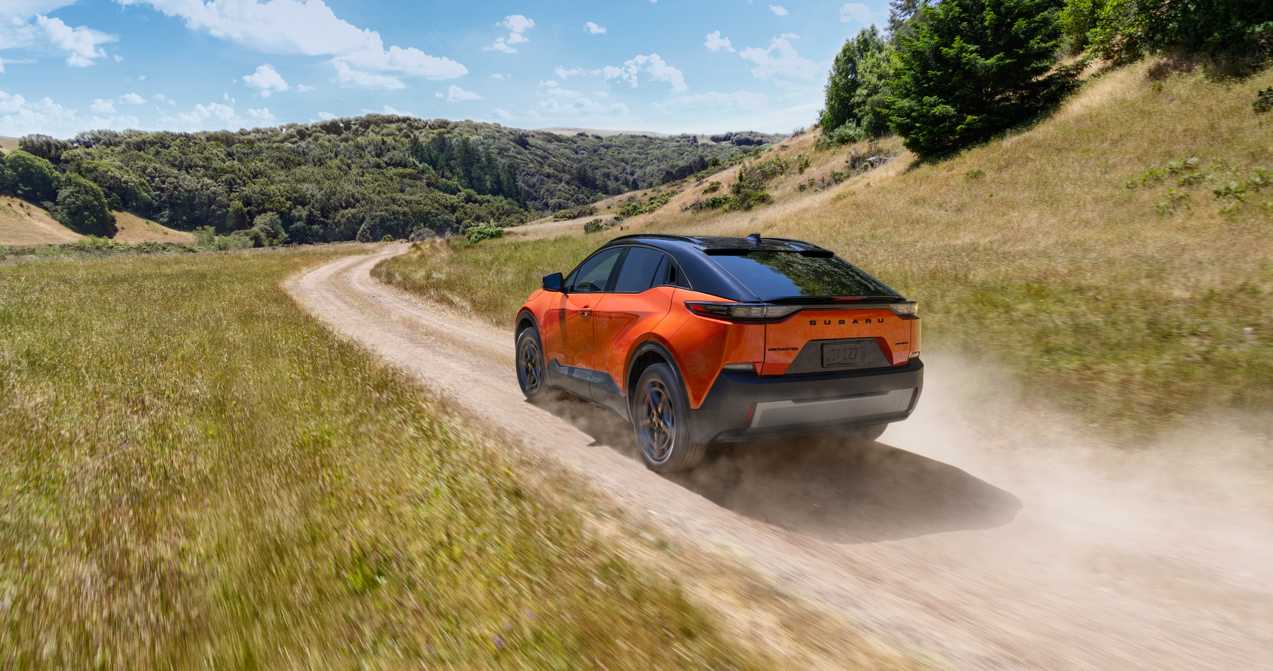 26_UNC_hero_gallery_1.png / An orange Subaru Uncharted electric SUV drives on a dirt road through some rolling hills.
