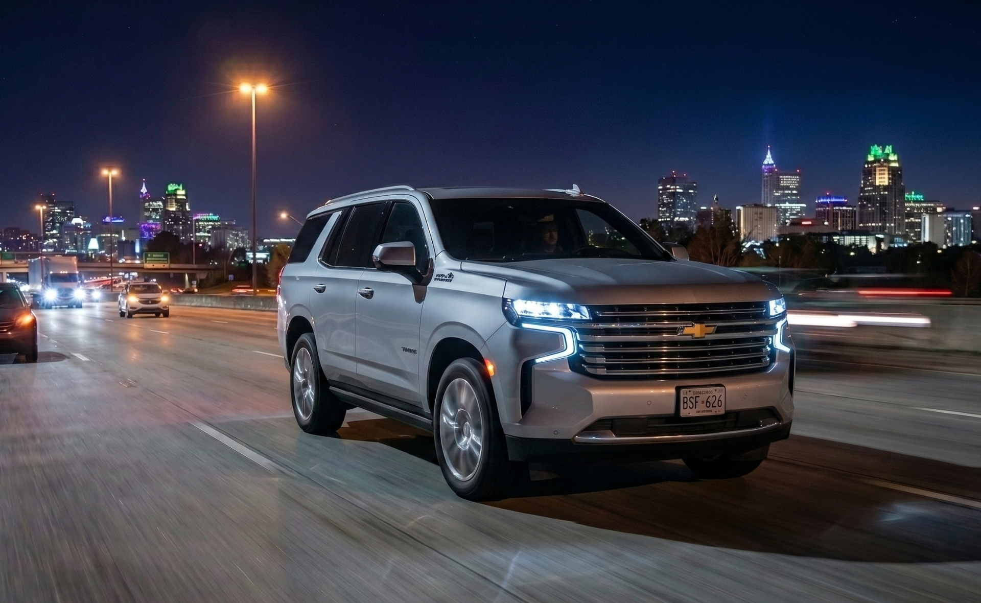 A silver Chevrolet Tahoe full-size SUV driving on a highway at night with the illuminated downtown Raleigh skyline, including the PNC Plaza spire, visible in the background.