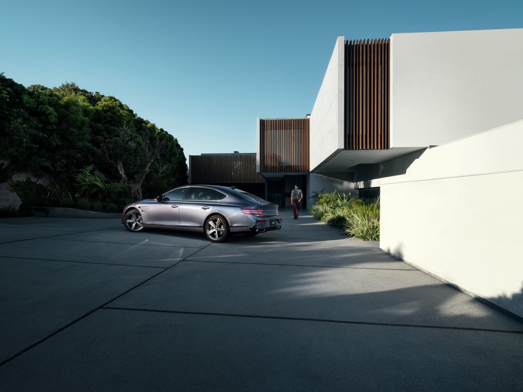 7/8 front driver side view of Genesis G80 in Ceres Blue parked next to modern house with man nearby