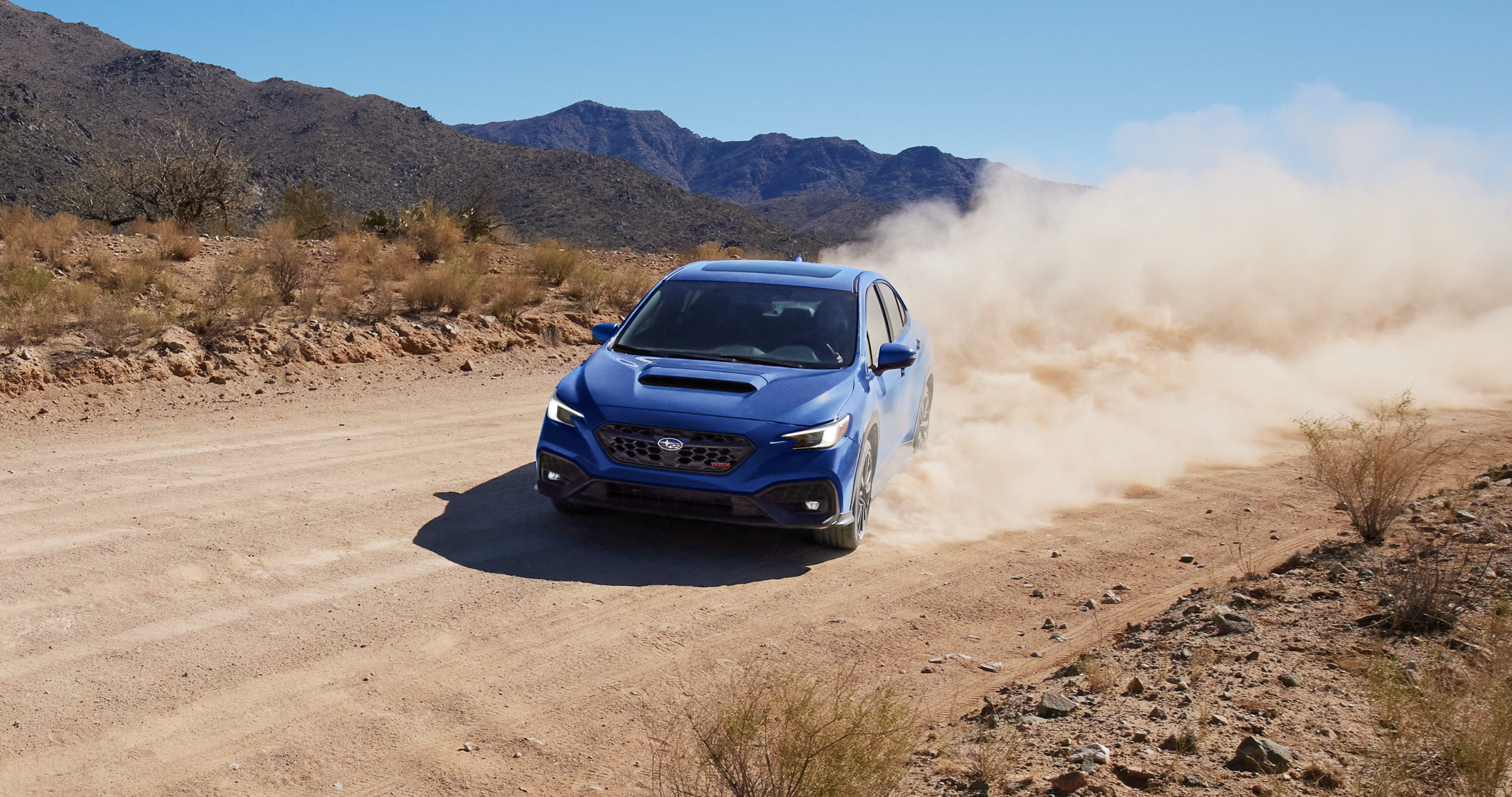 25_WRX_hero_gallery_1.png / A blue 2025 WRX 4-door sports car kicks up a large cloud of dust rounding a corner on a gravel road.