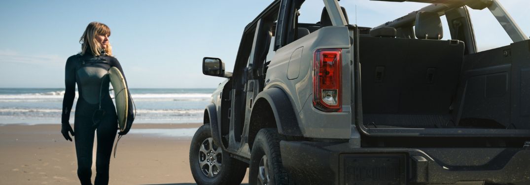 Woman with Surfboard Next to 2025 Ford Bronco with Open Tailgate at the Beach