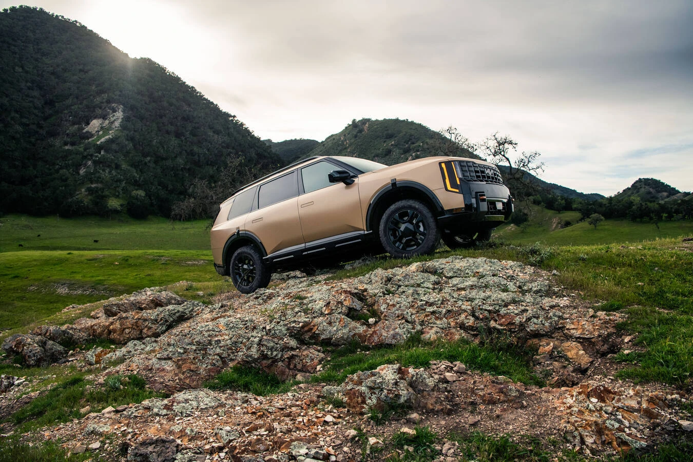 Bronze 2027 Kia Telluride X-Pro parked on a rocky outcrop, front three-quarter view, with green hills and mountains in the background.