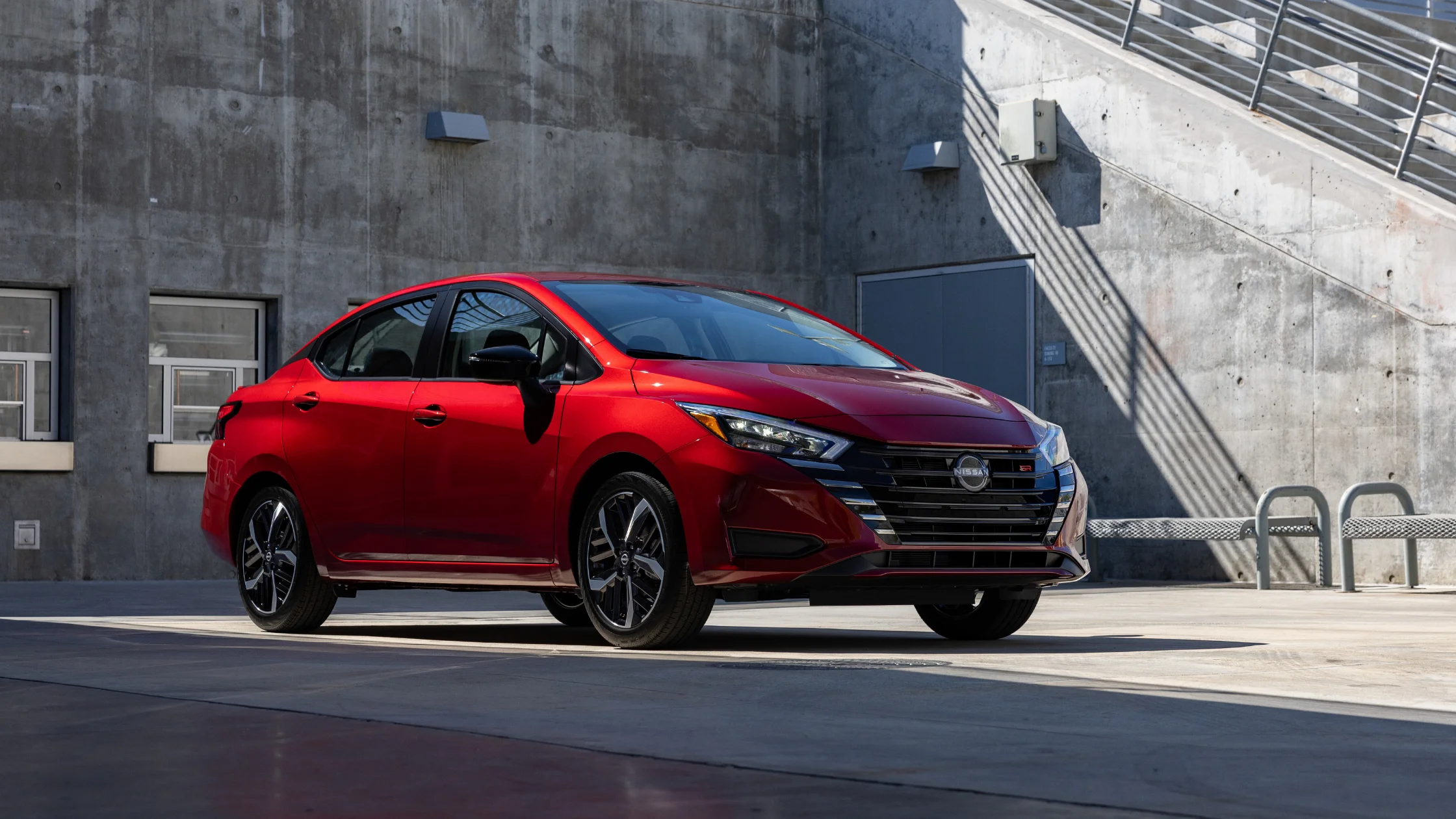 2025 Nissan Versa® in ruby parked outside a modern concrete building with sporty black alloy wheels.