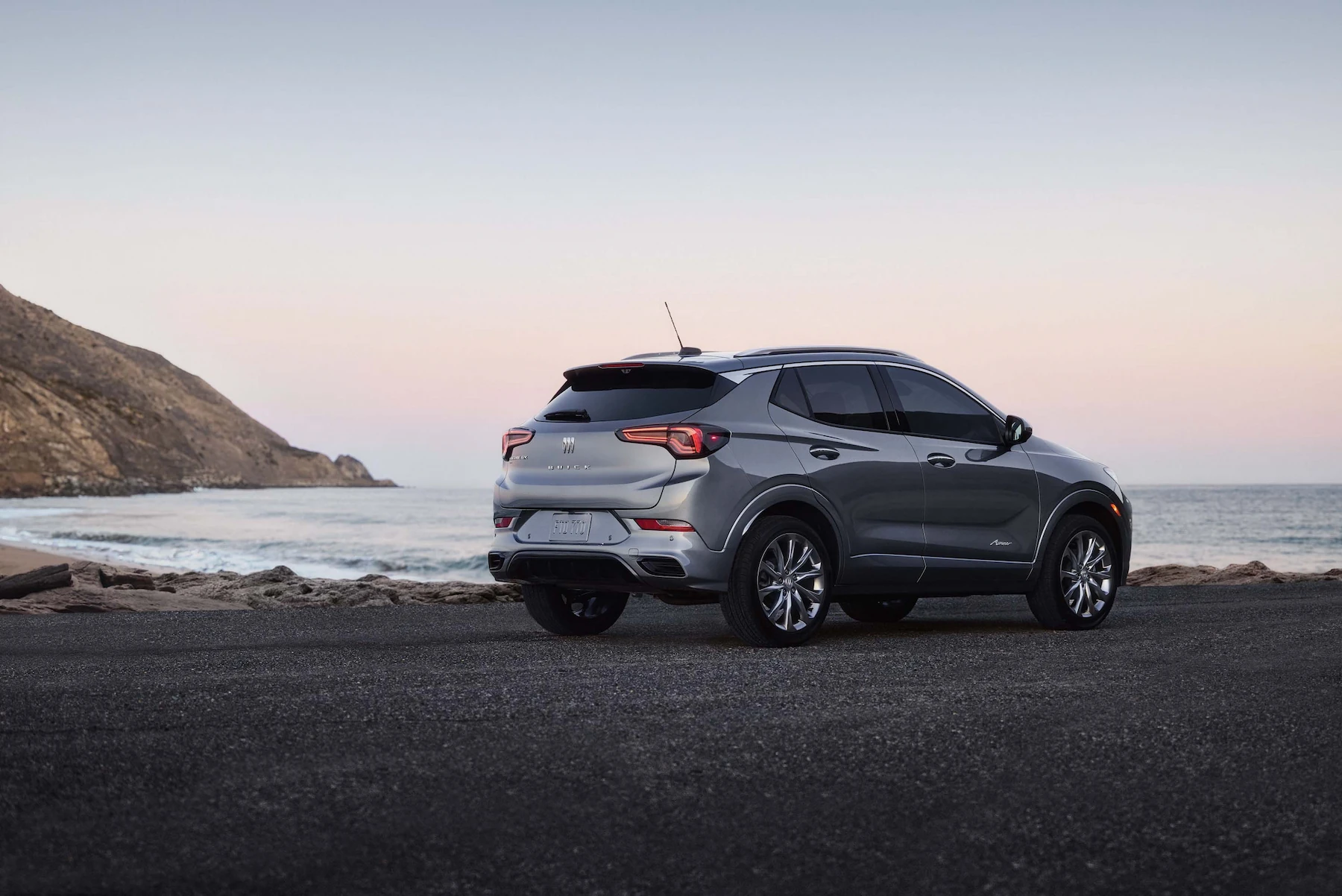 Rear three-quarter view of a silver Buick SUV with an Avenir trim badge, parked on a coastal road at dusk with the ocean and cliffs in the background