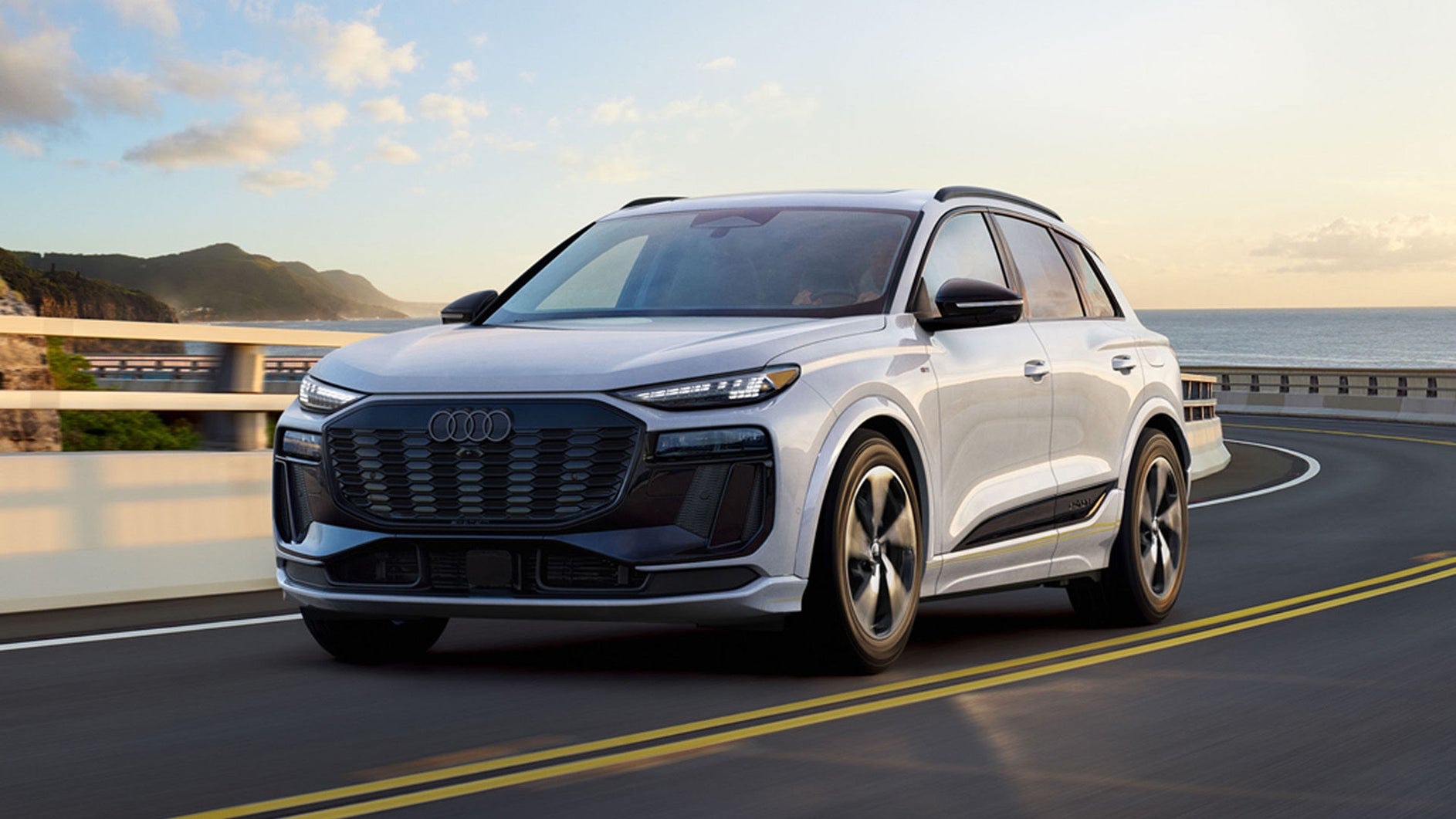 A white Audi Q6 e-tron drives along a coastal highway with ocean cliffs and a bridge in the background under low sunlight.