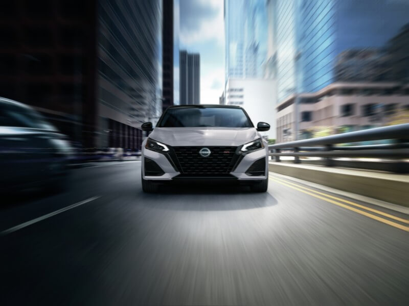 Front view of a silver 2026 Nissan Altima SR driving through a city at night with blurred buildings in the background