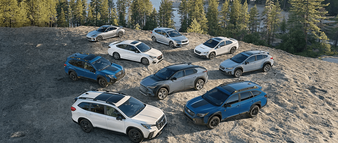 Subaru lineup of SUVs, wagons and sedans parked together on a rocky hilltop above a forest lake