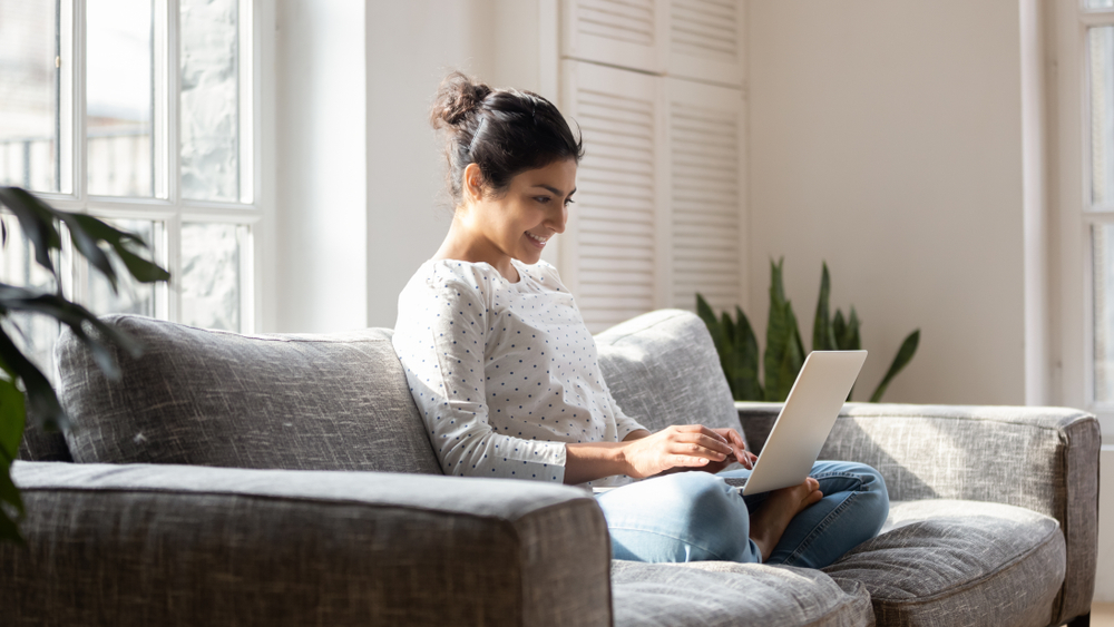 woman smiles as she sits on the couch with her laptop, researching used vehicles to buy