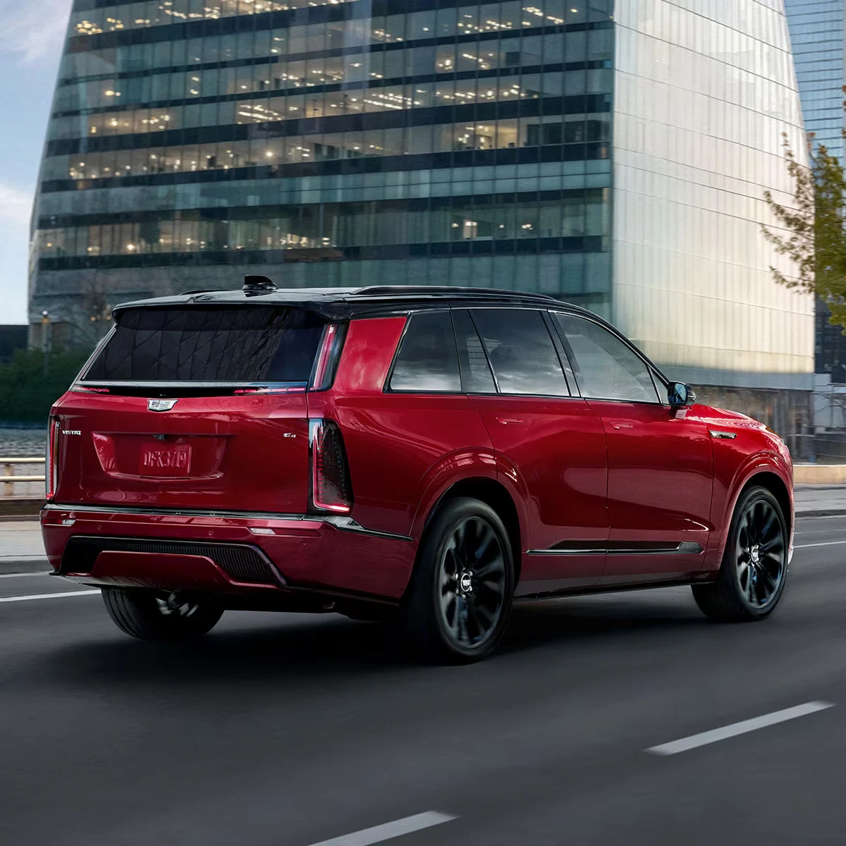 Red 2027 Cadillac Vistiq SUV with a black roof and black multi-spoke wheels, seen from the rear three-quarter driving along a city road, with a lit glass office building behind it