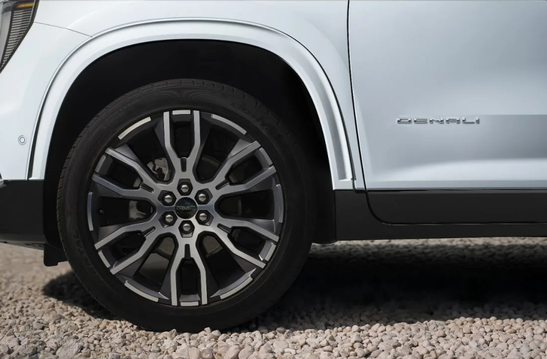 Close-up of a GMC SUV's front wheel and a legible "Denali" badge on the lower door panel, parked on a gravel surface