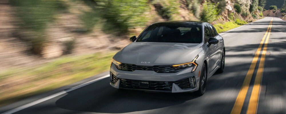 2025 Kia K5 driving on a scenic road, showcasing its sporty design and performance features.