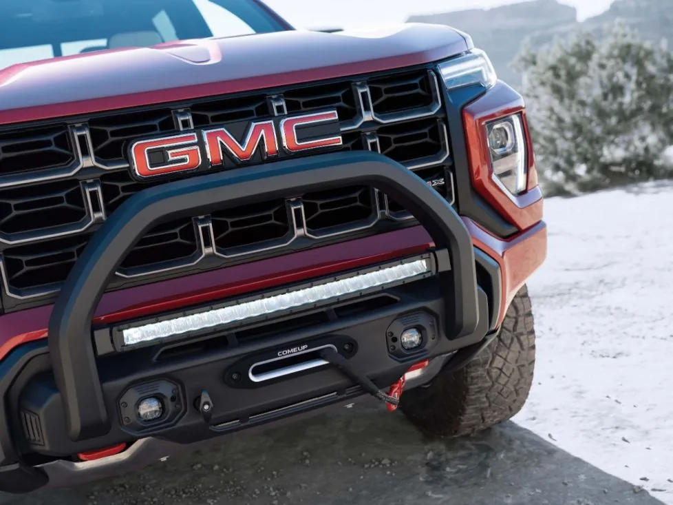 Close-up of a red GMC pickup truck's grille and front bumper, showing a legible red "GMC" wordmark, an off-road light bar, and a partial "AT4X" fender badge, with snow on the ground